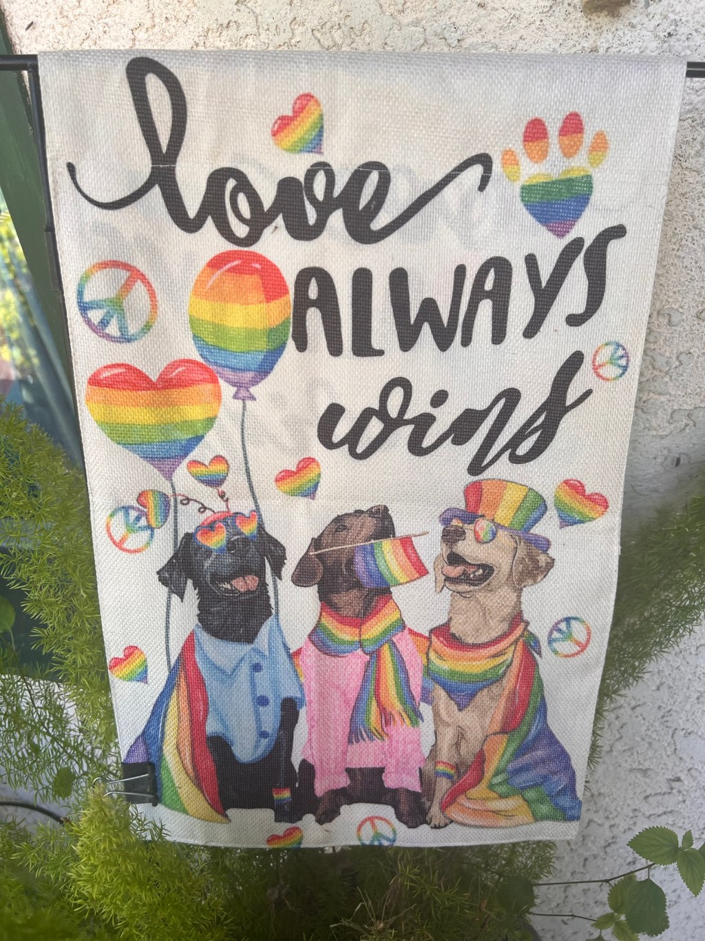 Flag reads in script Love Always Wins and features three dogs dressed in clothing. One is also wearing a rainbow hat. Two are wearing rainbow glasses. A couple have rainbow scarves one. There is a rainbow balloon of standard shape and one heart shaped. There are four rainbow peace signs scattered on the background.