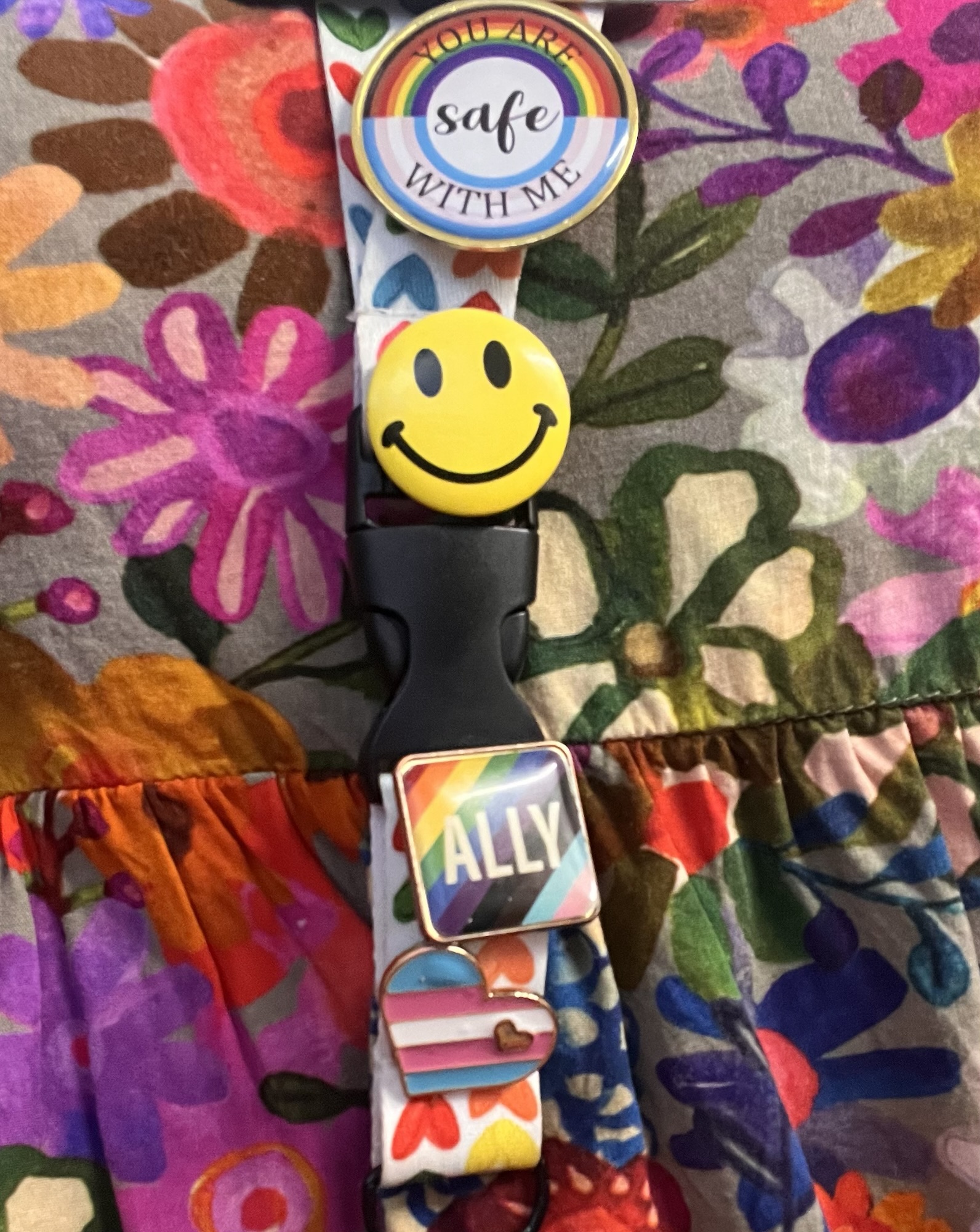 Cropped photo of 4 buttons appearing on a rainbow heart lanyard. Top pin read You Are Safe With Me, next down is a yellow smiley face, next down is a progressive rainbow background with the word ALLY in white and below that is a Transgender pride colored heart pin.