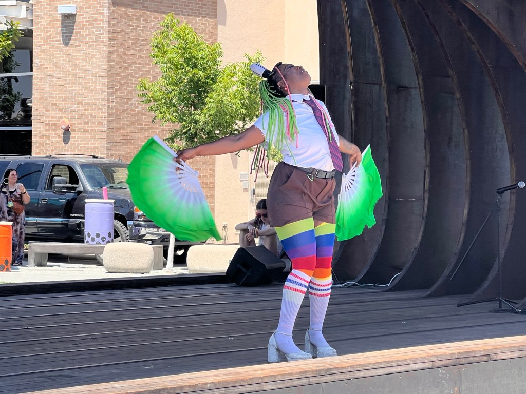 Drag performer on outdoor stage dancing with two larger green fans