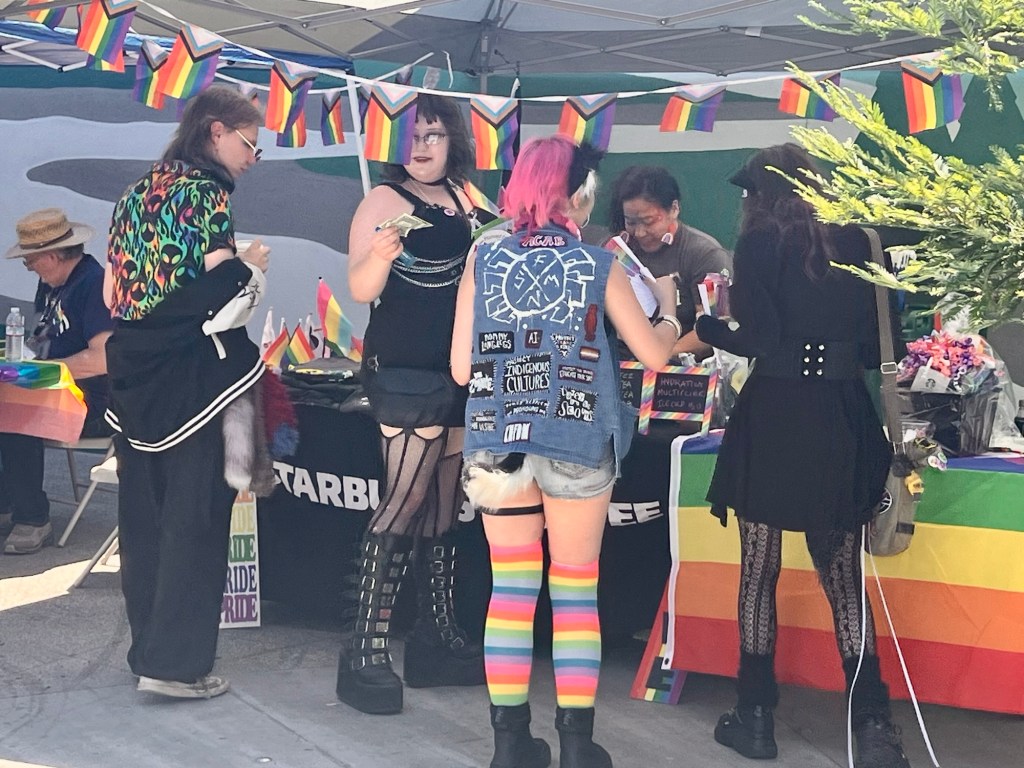 Young attendees at a Pride booth. They are wearing combos of black and colorful clothing and accessories