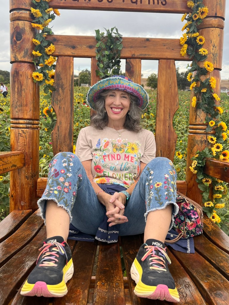 Author sitting in oversized wooden rocking chair in flower field. She is wearing flower embroidered jean capris, a flower tshirt that reads Find Me In the Flowers and a colorful brimmed hat. 